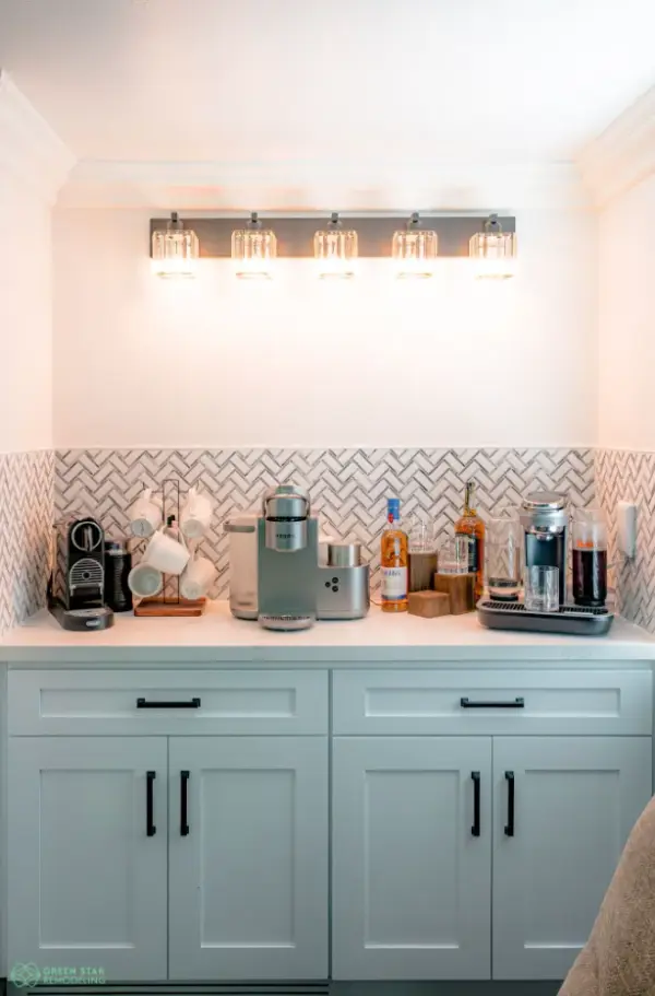 A modern home coffee and beverage bar featuring light gray shaker cabinets with black hardware, a white herringbone tile backsplash, and a five-light crystal vanity fixture.