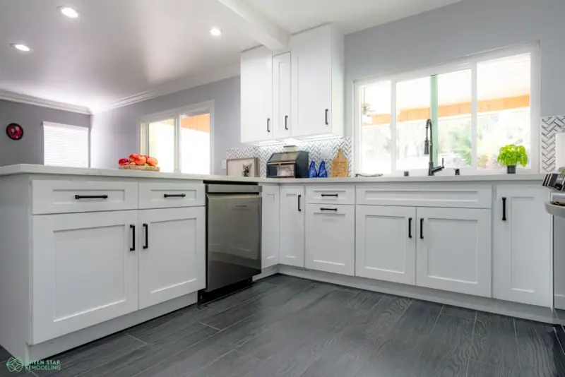 A modern kitchen remodel featuring white shaker-style cabinets with black hardware, a light gray herringbone tile backsplash, and dark wood-look flooring.