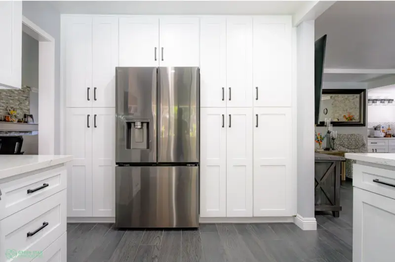 A modern kitchen remodeling project in Santa Clarita featuring floor-to-ceiling white shaker cabinets with black hardware surrounding a stainless steel French door refrigerator on dark wood-look tile flooring.
