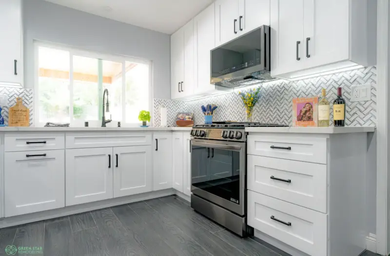 Modern kitchen remodel featuring white shaker cabinets with black hardware, a herringbone tile backsplash, stainless steel appliances, and dark wood-look flooring.