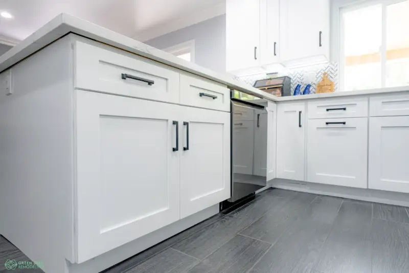 A bright modern kitchen featuring white shaker-style cabinets with black hardware, white quartz countertops, and dark grey wood-look tile flooring.