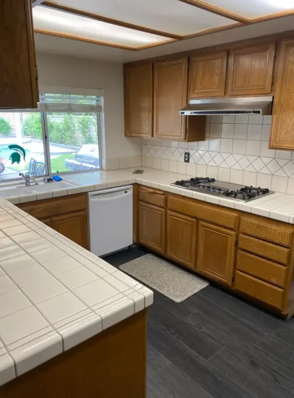 A residential kitchen featuring traditional honey oak wood cabinetry, white square tile countertops with matching backsplash, a built-in gas stovetop under a stainless steel range hood, and a white dishwasher.