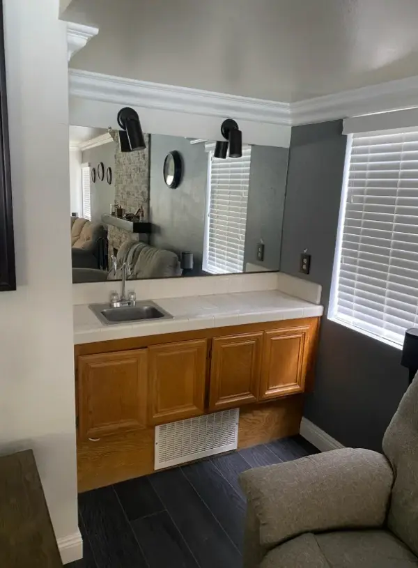 A modern home wet bar featuring a white tiled countertop with a small stainless steel sink, oak wood cabinetry, and a large mirror reflecting a living area with a brick fireplace.