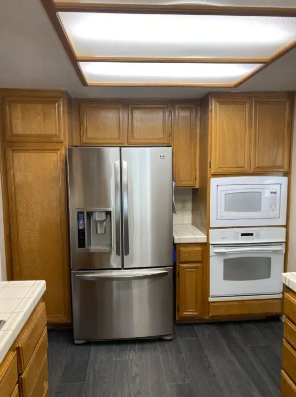 A modern stainless steel French door refrigerator integrated into traditional oak wood cabinetry with a built-in white microwave and oven.