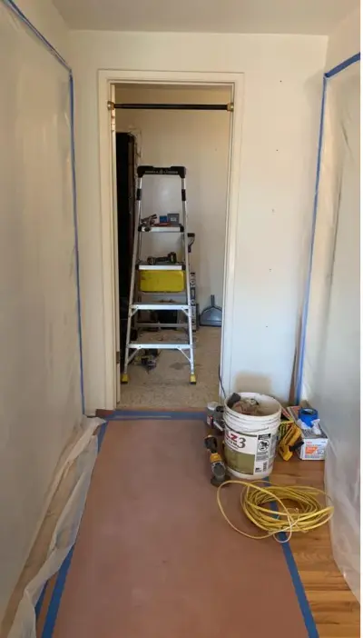 Bathroom renovation in progress showing construction tools, protective plastic sheeting, a ladder, joint compound bucket, and power cord inside a remodeled bathroom space.
