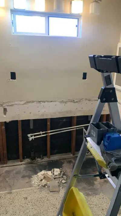 A bathroom undergoing renovation with an open wall section exposing wooden studs and plumbing pipes, featuring a stepladder and construction debris in the foreground.