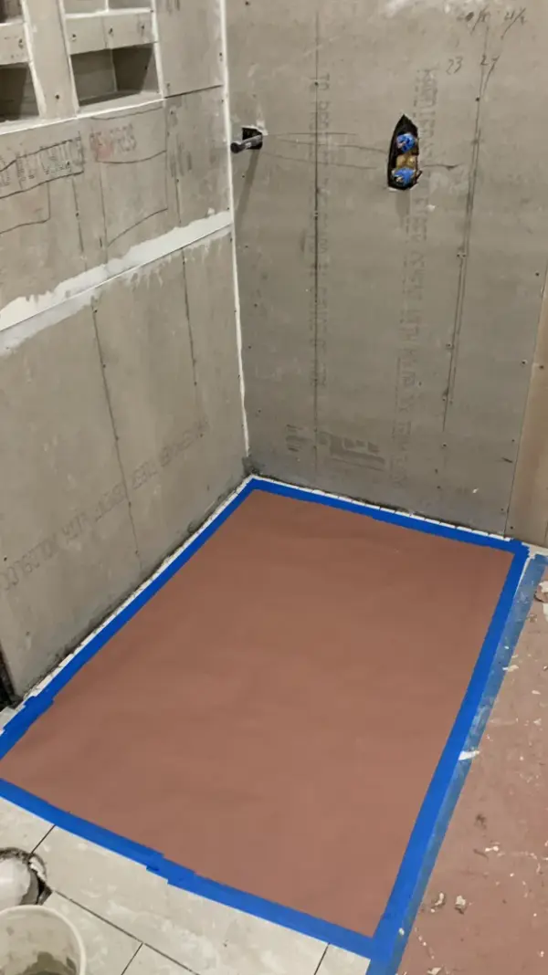 A bathroom shower under renovation featuring installed cement backer board on the walls and a protected floor area taped off with blue painter's tape.