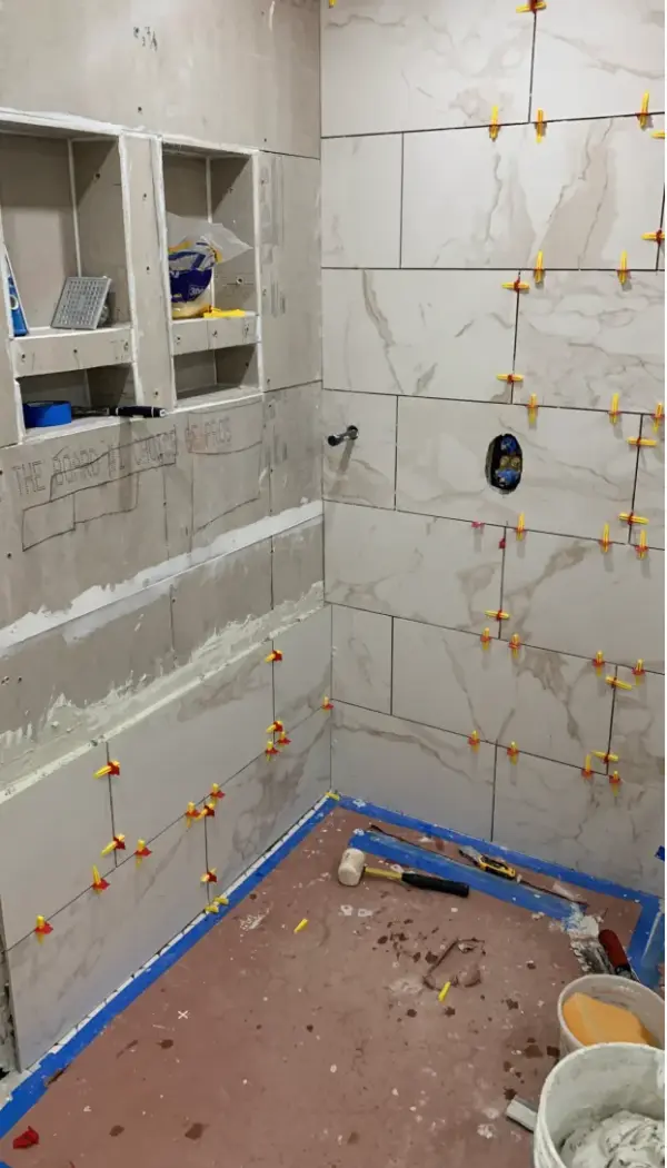 A bathroom shower under renovation featuring large marble-patterned wall tiles being installed with a yellow leveling system, built-in wall niches, and various tiling tools on the floor.