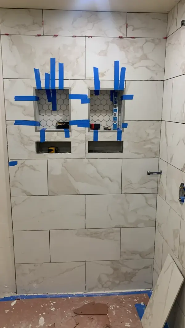 A bathroom remodeling project showing the professional installation of large-format white marble tiles on a shower wall featuring two built-in storage niches with blue painter's tape and leveling tools.