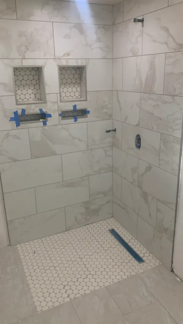 A modern walk-in shower under renovation featuring white marble-style wall tiles, two built-in niches with circular penny tile accents, and a matching penny tile shower floor with a linear drain.