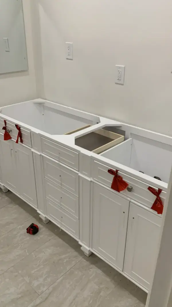 A white double-sink bathroom vanity being installed against a light wall, featuring recessed panel cabinets and drawers with protective red ribbons on the hardware.