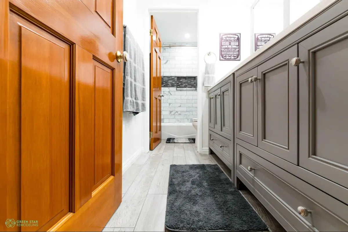 Modern renovated bathroom featuring custom grey double vanity cabinets with brushed nickel knobs, light grey wood-look tile flooring, and warm natural wood doors. The en-suite opens into a tiled shower and bath area lined with classic white subway tile and a dark mosaic accent niche.