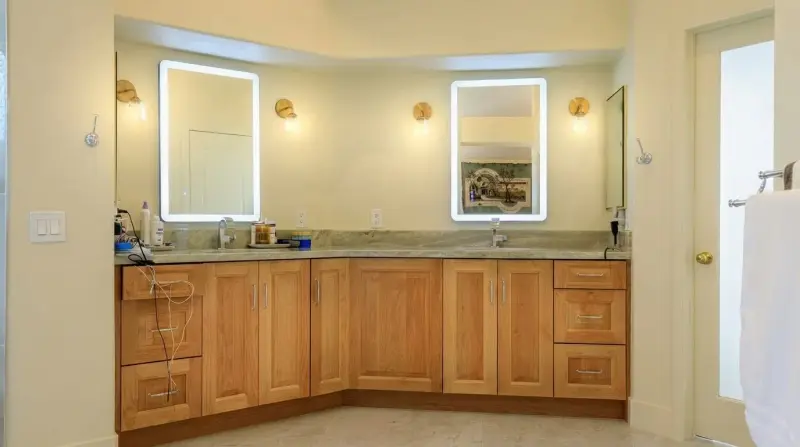 Double sink vanity with wood cabinetry, backlit LED mirrors, and cream-colored stone countertops.