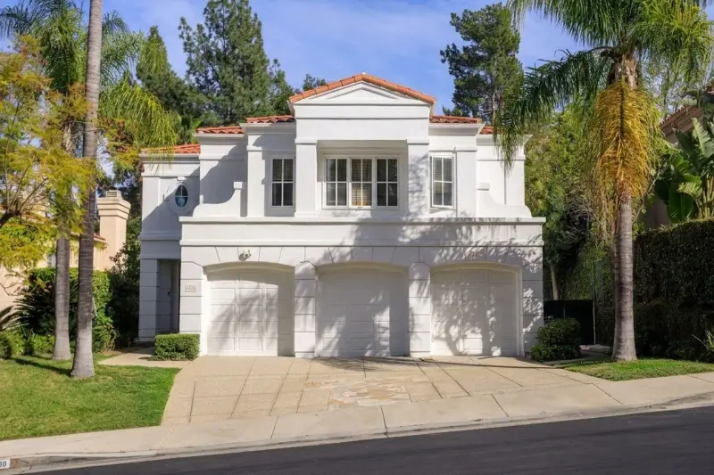 Exterior view of the two-story white stucco home featuring Mediterranean architecture, a red tile roof, three-car garage, and palm tree landscaping.
