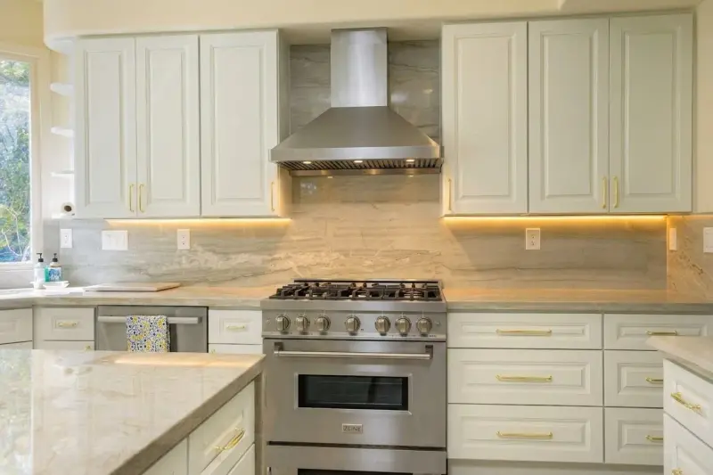 Kitchen detail showing upper and lower white cabinets with brushed gold handles, stainless steel range hood, and natural stone backsplash.