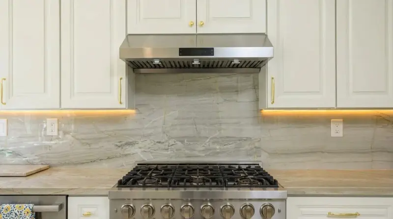 Close-up of the stainless steel gas range, full-height natural stone backsplash, and white cabinetry with brushed gold hardware.