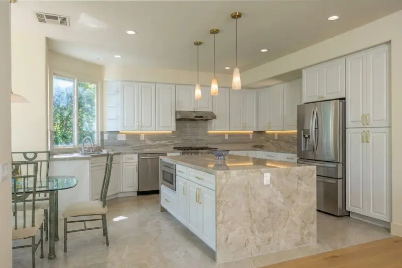Kitchen layout showing the central stone island with waterfall edges, surrounding white raised-panel cabinetry, and gold hardware.
