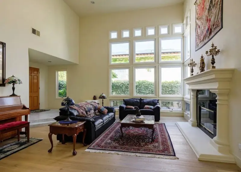 Spacious living room with high ceilings, dark leather sofas, a traditional stone fireplace, piano, and large grid windows.