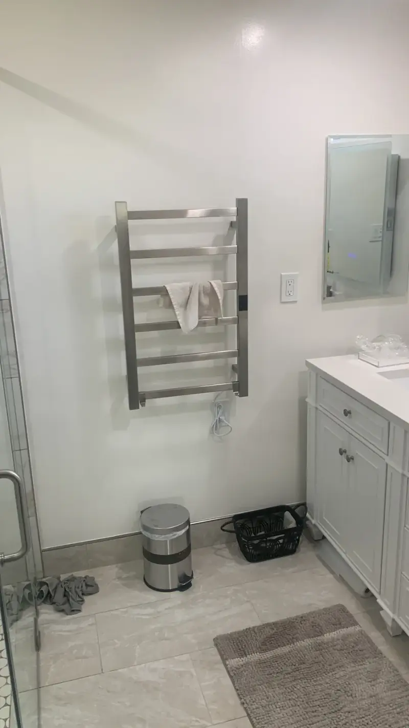 A modern bathroom interior featuring a wall-mounted stainless steel heated towel rack with a white cloth, positioned next to a white vanity cabinet and a mirror.