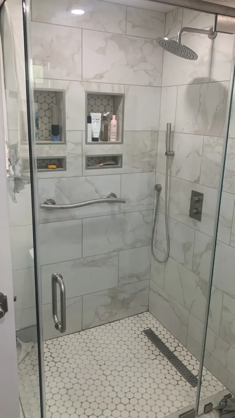 A modern walk-in glass shower featuring white marble wall tiles, a penny tile floor with a linear drain, and four built-in storage niches holding toiletries.