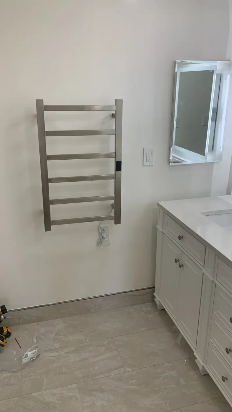 A modern brushed metal wall-mounted electric towel warmer installed on a white bathroom wall next to a vanity and mirror.