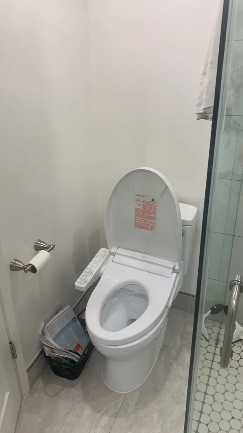 A modern white toilet with an open lid and a bidet control panel attached to the side, situated in a brightly lit bathroom.