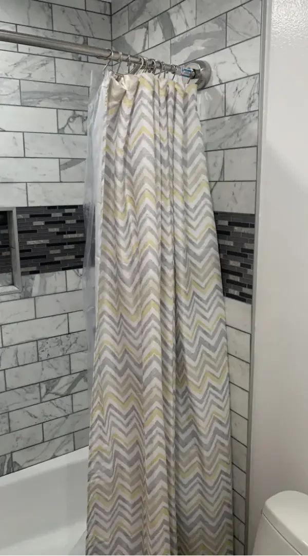 A modern shower curtain featuring a gray and yellow chevron zigzag pattern hanging in a bathroom with marble-style subway tiles and a dark accent tile border.